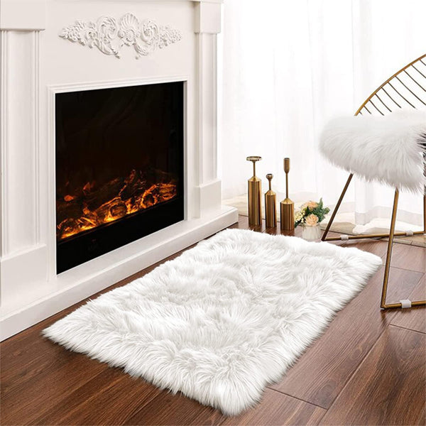 The Latepis White Luxury Faux Fur Sheepskin Rug 2x3 is placed on a wooden floor near a lit fireplace, accompanied by a gold chair and elegant decor items.